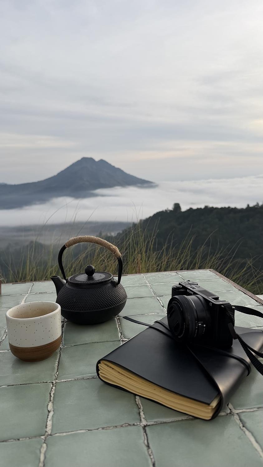 A teapot, a cup, a notebook and a camera on a tiled table on the caldera rim, the volcano in the mist behind