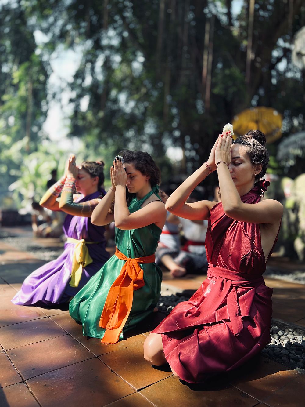 Three women in sarongs kneeling in prayer, palms together, at Taman Beji Griya