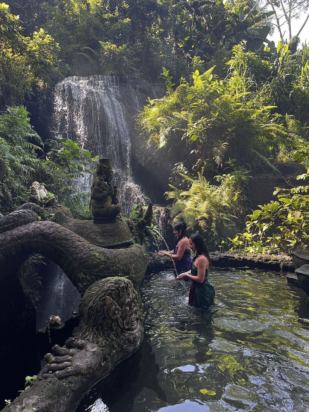 The waterfall pool at Taman Beji Griya temple, a carved dragon on the rocks, two women in the water