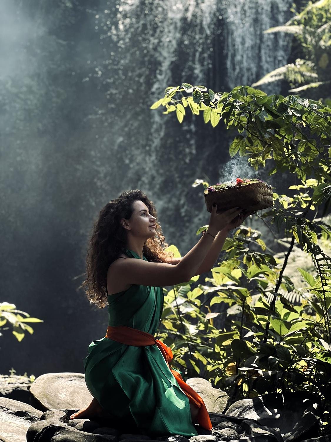 A woman in a green sarong kneeling on the rocks at Taman Beji Griya, holding an offering up into the spray of the waterfall