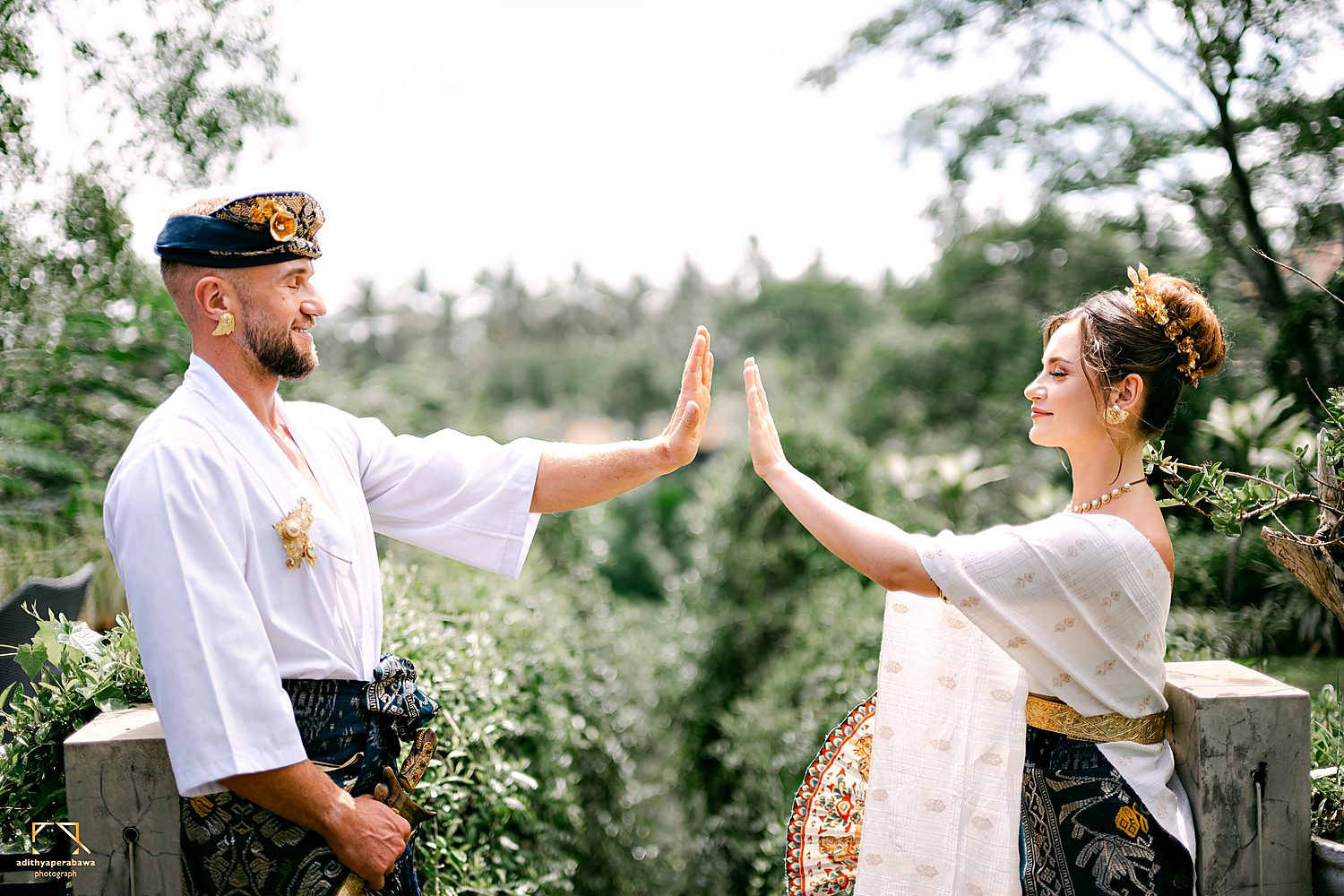 A couple in traditional Balinese dress, palms touching, the jungle behind them