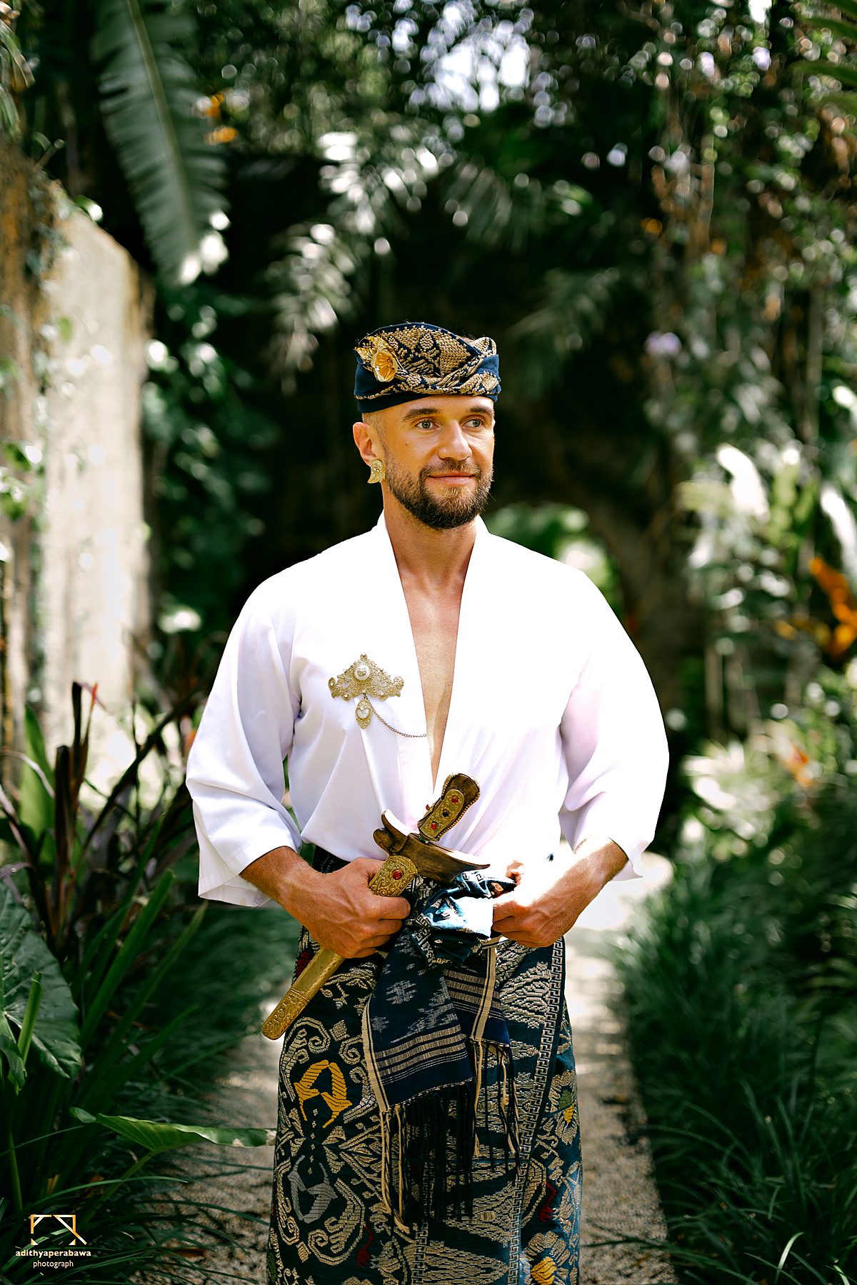 A man in a Balinese udeng headdress and white shirt, a kris at his belt, in the garden