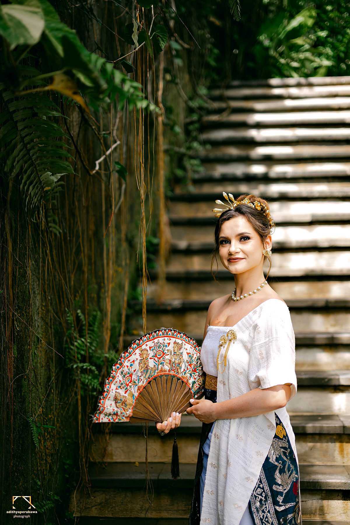 A woman in a white kebaya with a painted fan, by the stone steps