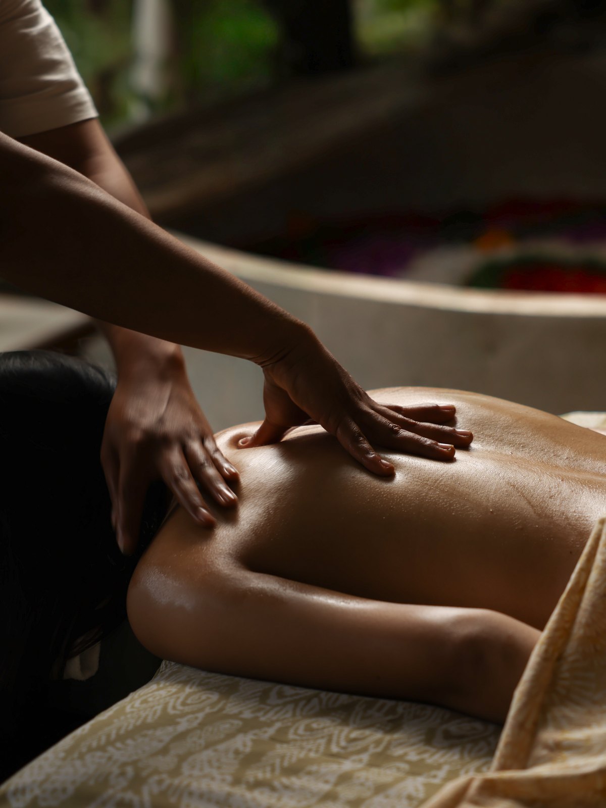 A therapist&rsquo;s hands on a guest&rsquo;s back, the flower bath behind