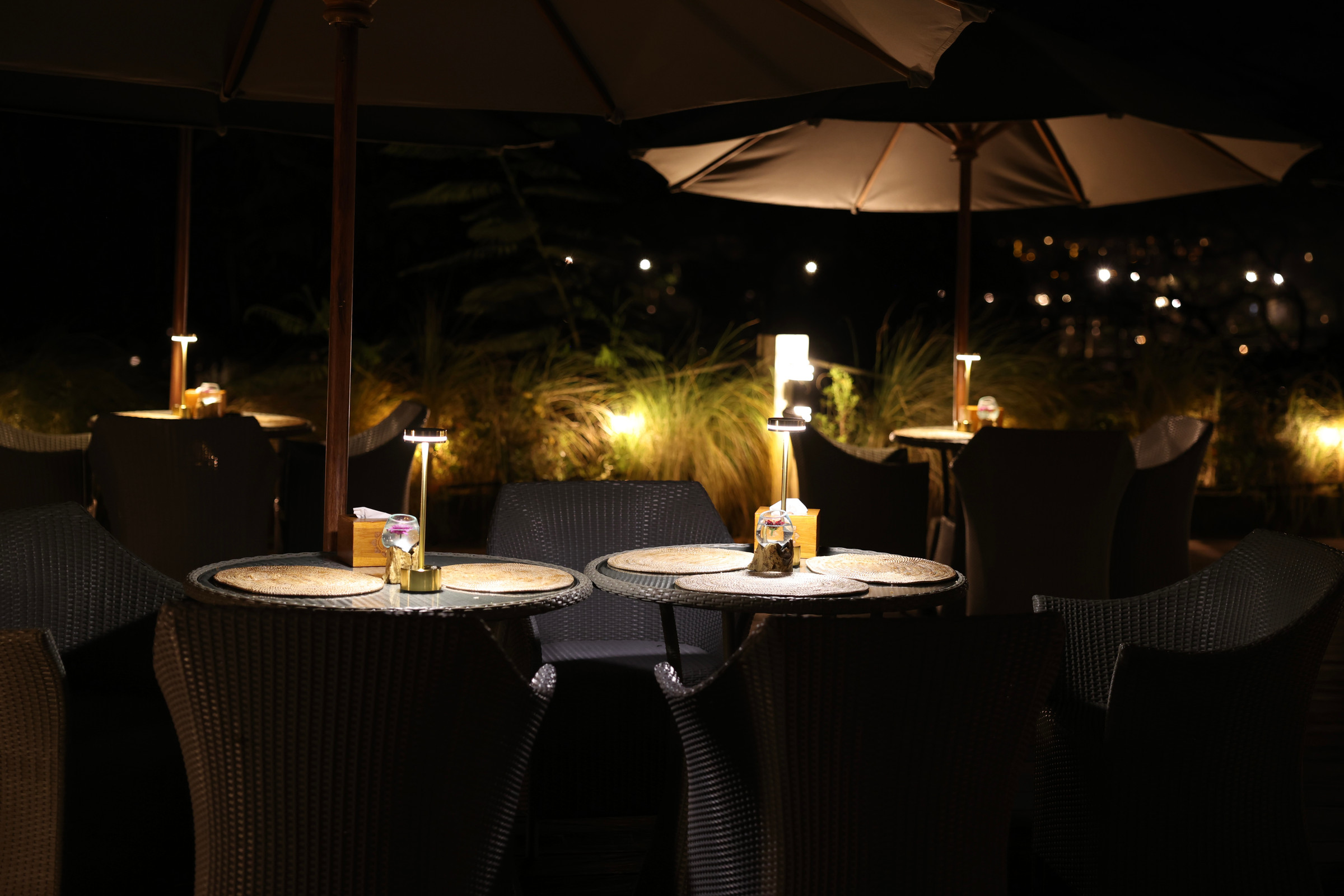 Tables under the umbrellas on the terrace at night, a lamp on each and the lights of the far side of the valley behind