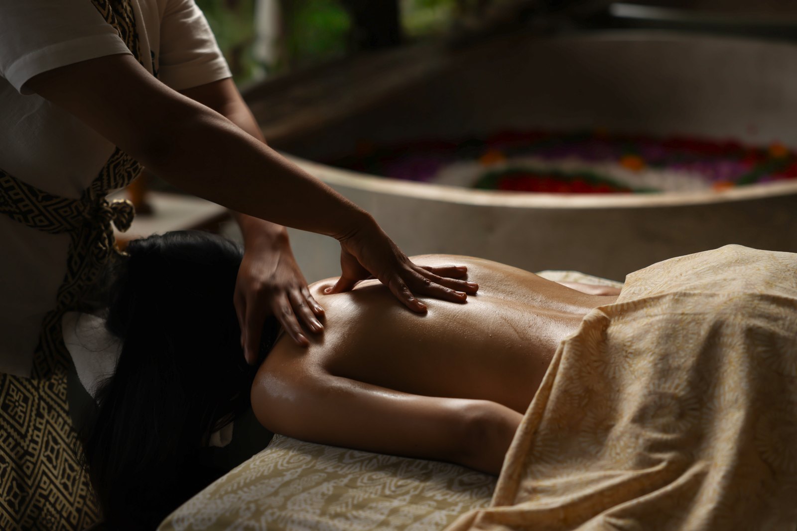 A Balinese massage: the therapist&rsquo;s hands on the back, warm oil, the flower bath behind